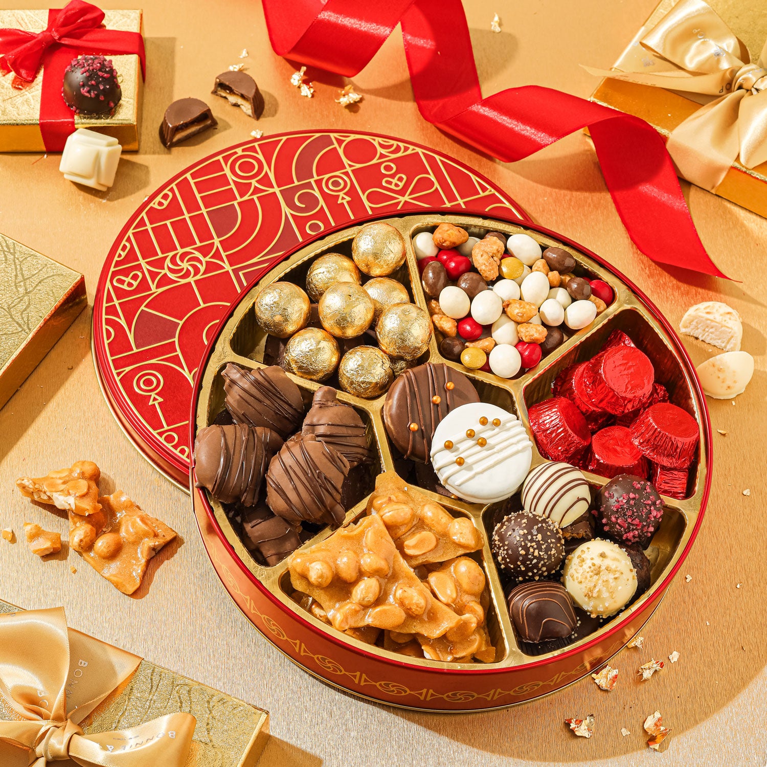 A festive red and gold circular box of assorted gourmet sweets including chocolate covered treats, nut clusters, and candy balls.