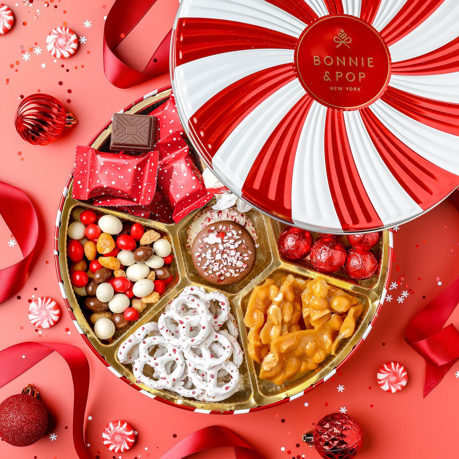 A red and white tin with a Lazy Susan label, filled with assorted holiday treats including peanuts, chocolate covered treats, Oreos, taffy, and pretzels, all arranged in sections.