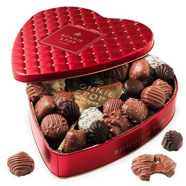 A red heart-shaped tin filled with an assortment of chocolates and chocolate-covered snacks, including truffles and pretzels, with a decorative pattern on the lid.
