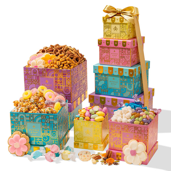Summer Gift Tower, Gourmet Candy & Snack Box Assortment with Popcorn, Cookies, Taffy, Nuts, Caramels & Sweet Treats - Bonnie & Pop. This gourmet gift tower features a collection of stacked, square-shaped cardboard boxes in vibrant shades of purple, teal,
