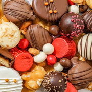A festive red and gold circular box of assorted gourmet sweets including chocolate covered treats, nut clusters, and candy balls.